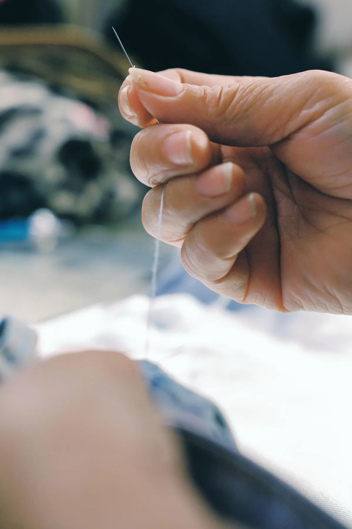 Close-up of hands threading fine cotton yarn through a sewing needle representing premium denim craftsmanship